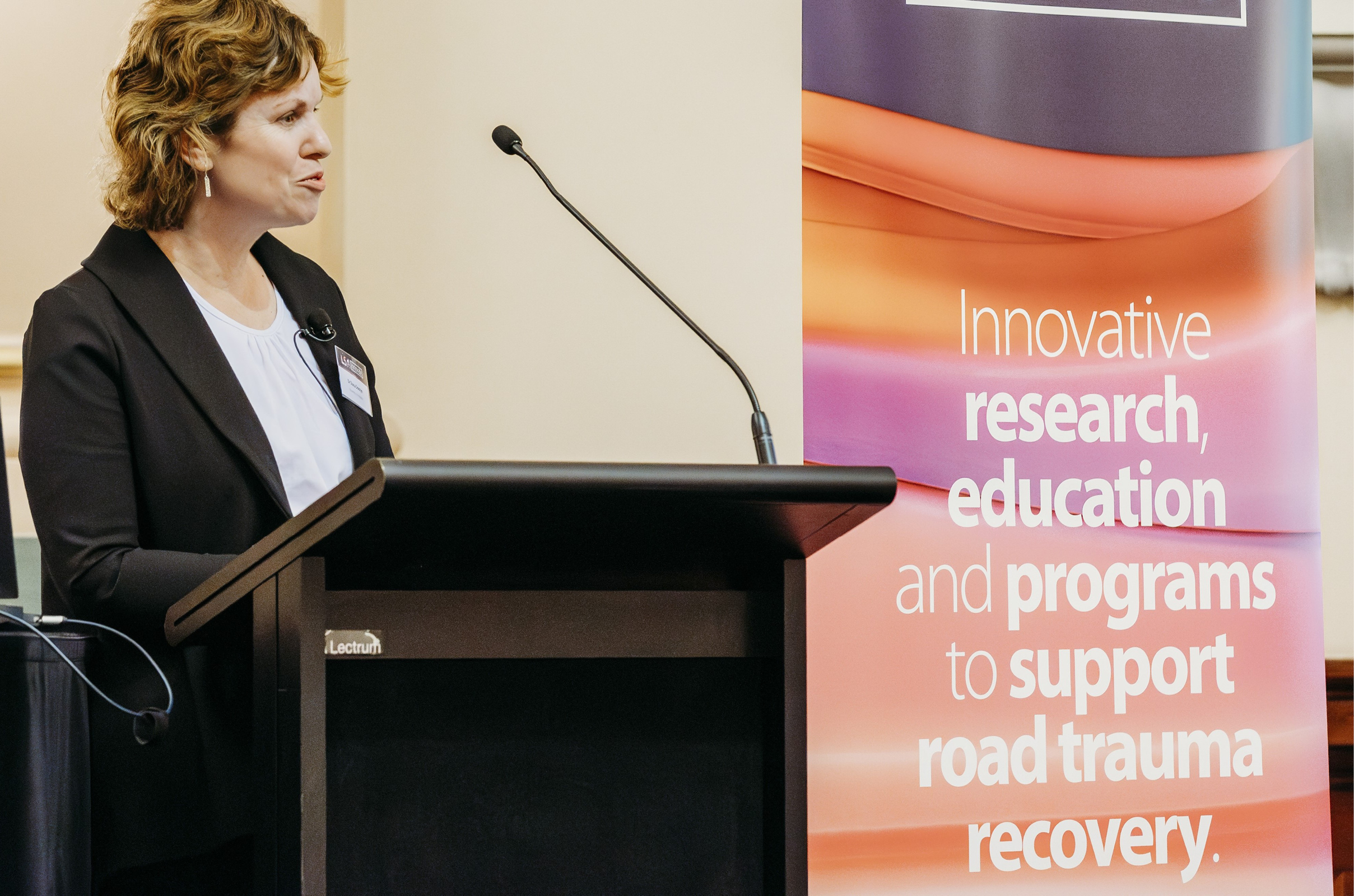 Woman speaking at a lectern in front of a banner which says innovative research, education and programs to support road trauma recovery